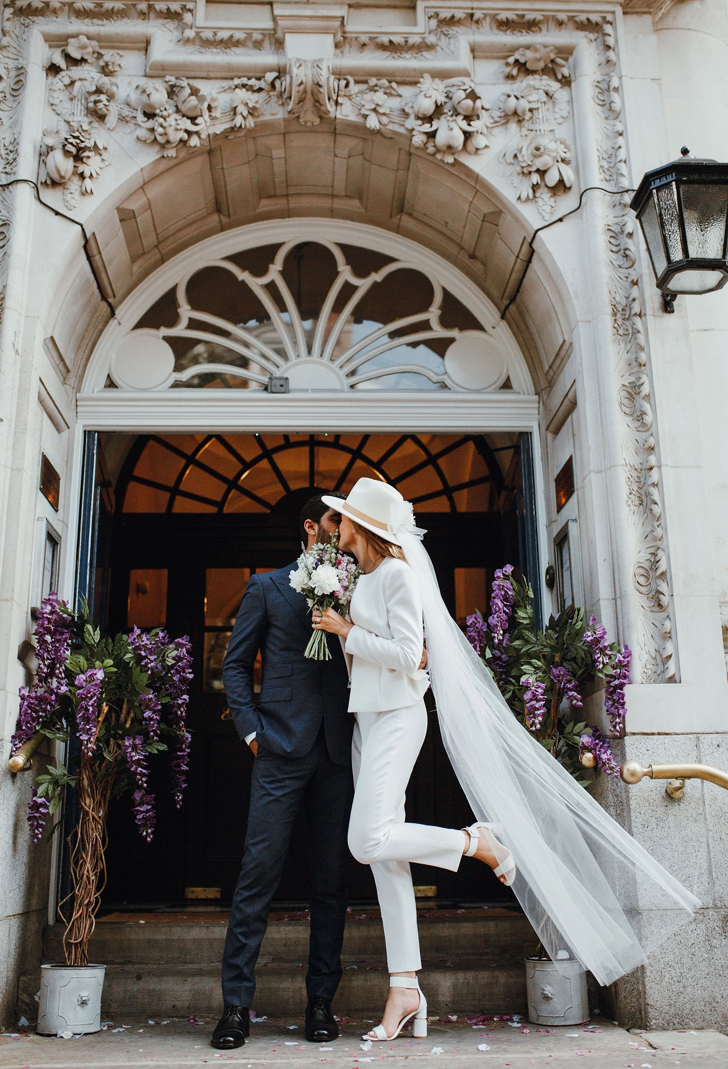 bride wears a white wedding suit and her groom in front of a church