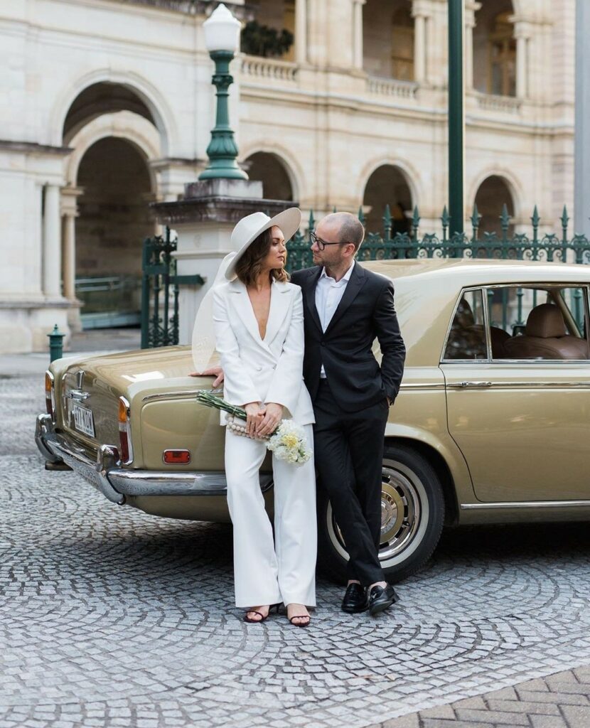 bride in a white wedding suit with groom in front of a classic luxury car