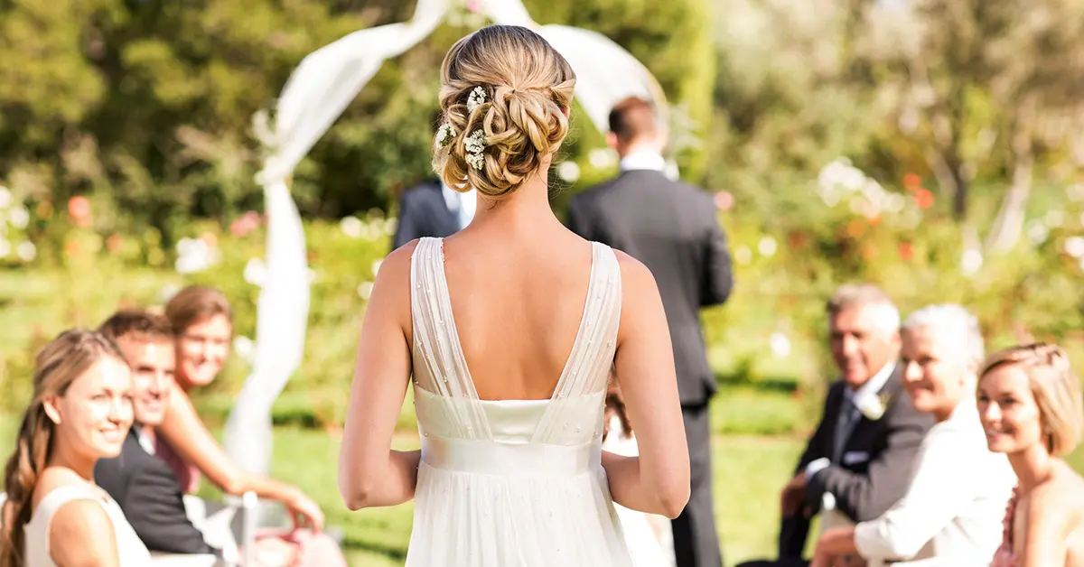 Bride walking down outdoor wedding aisle toward ceremony arch with seated guests